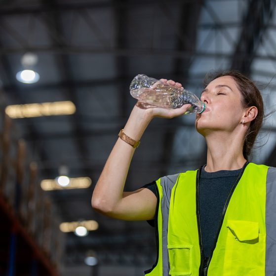 A woman in a high visibility vest drinks water from a plastic bottle inside a warehouse with metal shelves and a high ceiling.