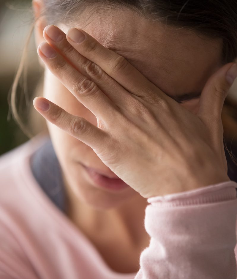 A woman with her hand covering her face, appearing distressed or upset. She has brown hair pulled back and is wearing a light pink top.