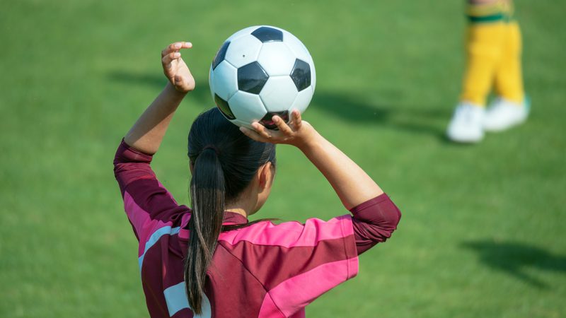 A girl in a pink and maroon sports jersey prepares to throw a soccer ball over her head during a game on a grassy field.