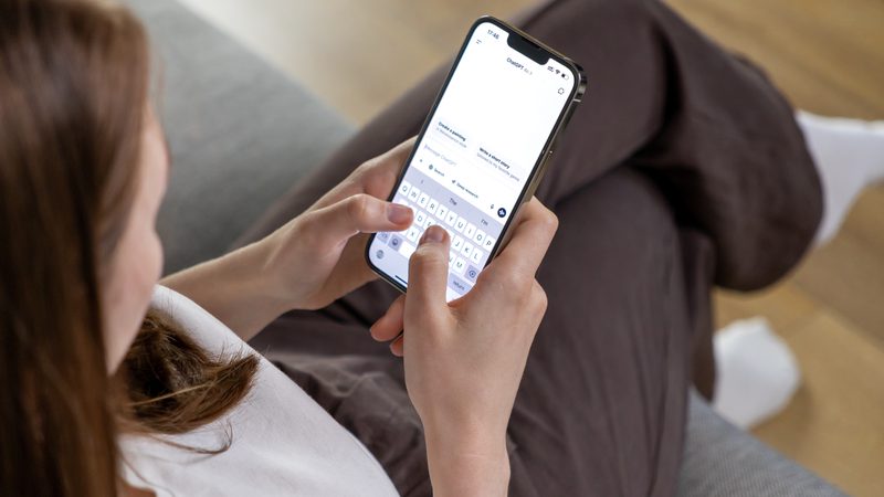 A person with long hair wearing a white top is sitting on a grey couch, holding a smartphone and typing a message.