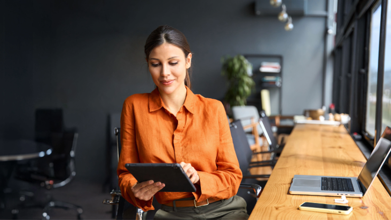 Person in an orange shirt sits at a long wooden table in a modern workspace, using a tablet while a laptop and smartphone rest nearby.