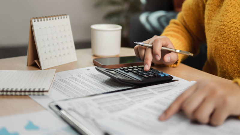 Person in a yellow sweater using a calculator and pointing at papers, with a notepad, coffee mug, smartphone, and calendar on the desk.