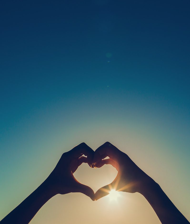 Two hands form a heart shape around the sun against a clear sky, with a tree visible on the right side. The sunlight shines through the heart shape.