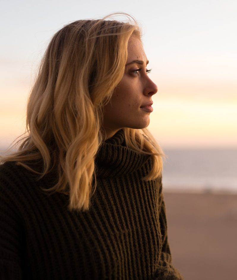 Profile of a blonde-haired individual in a dark chunky knit turtleneck, standing on a beach at sunset and gazing towards the horizon as the sea glows softly.