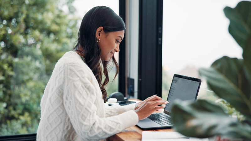Woman in a white sweater working on a laptop at a wooden desk by a window with green trees outside.