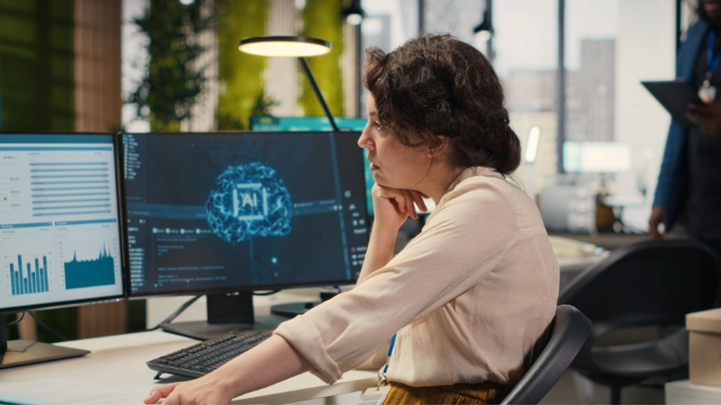 Person seated at a desk in a modern office, examining multiple monitors showing charts and a brain graphic, with hand on their chin.