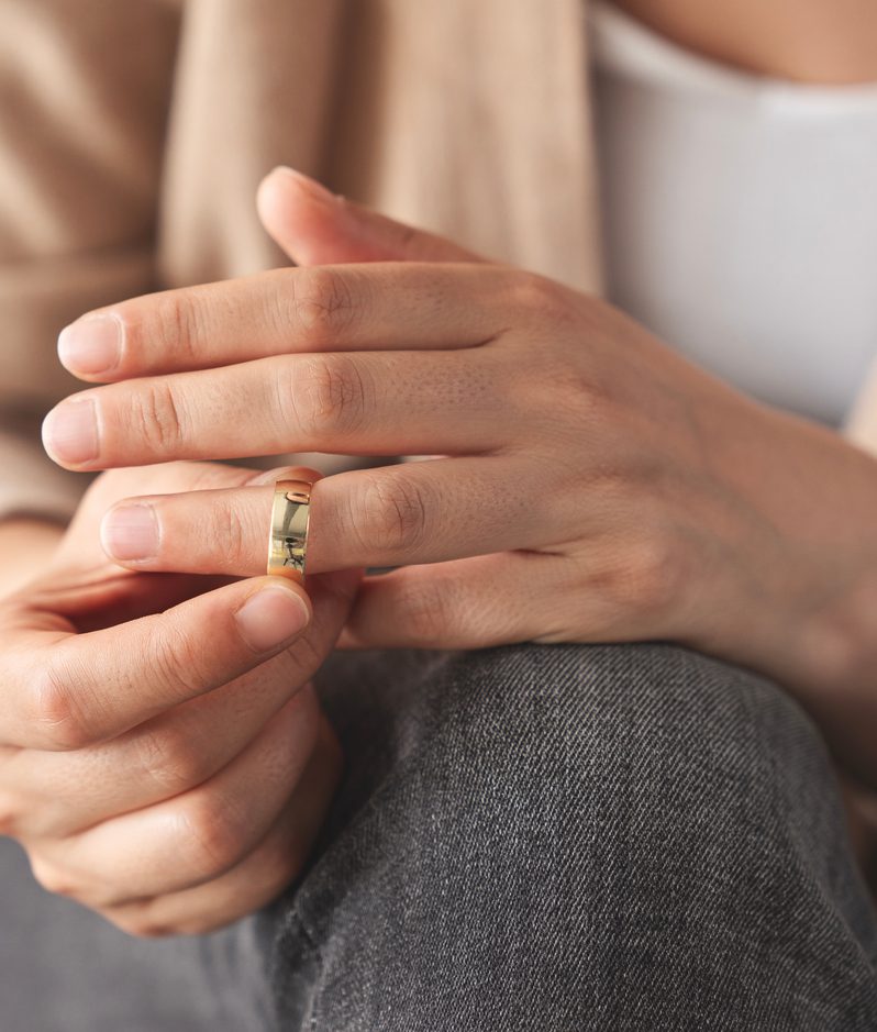 Close-up of hands with a gold ring on the ring finger, resting on a knee; the person wears a beige cardigan and denim jeans in a relaxed pose.
