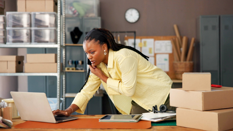A woman in a yellow shirt looks concerned as she observes her laptop screen in a storage or warehouse space surrounded by boxes, files, and office supplies.