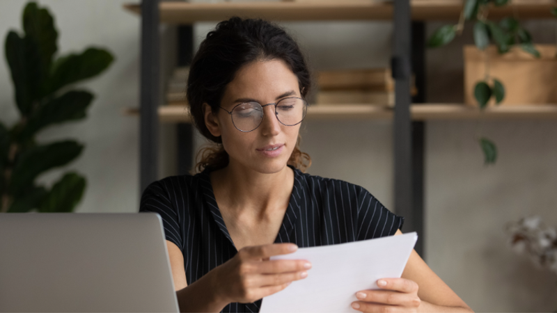 Person with glasses sits at a desk, examining a sheet of paper beside a laptop; a striped top, an indoor workspace with shelves and leafy plants in the background.