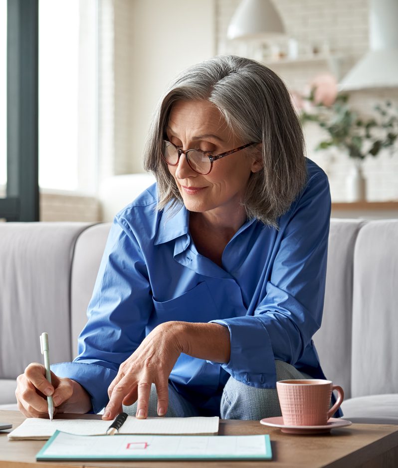 An older person with grey hair and glasses sits on a sofa in a bright living room, leaning over a notebook with a pen beside a laptop and a pink mug.