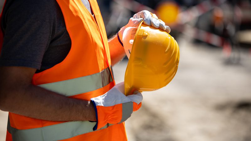 A construction worker wearing an orange safety vest and gloves holding a yellow safety helmet at a construction site.