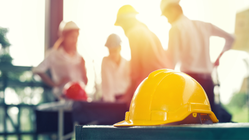 Yellow safety helmet rests on a metal surface in the foreground, while a sunlit group of workers in hard hats discuss plans at a construction site.