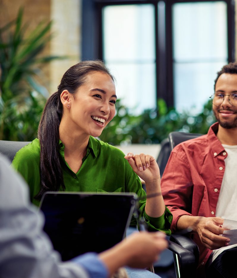 A group of four young adults engaged in a discussion in a modern, bright office space with large windows and exposed brick walls.