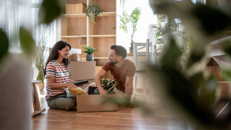 A man and woman sitting on the floor of a bright, modern room with wooden shelves and large windows, opening a box of houseplants and smiling at each other.