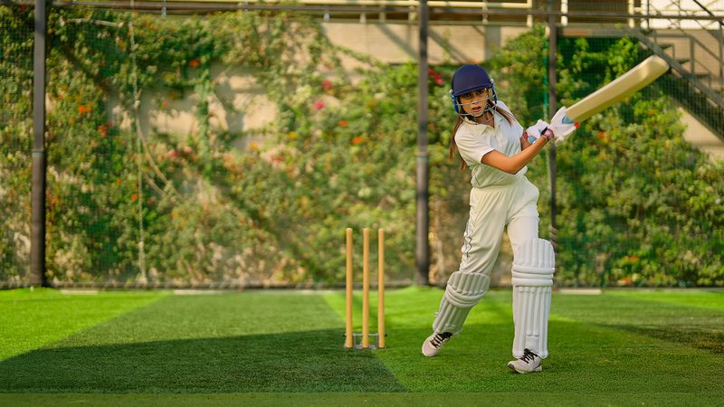 A girl in cricket whites, wearing a blue helmet, batting with a cricket bat on a green cricket field with stumps behind her.