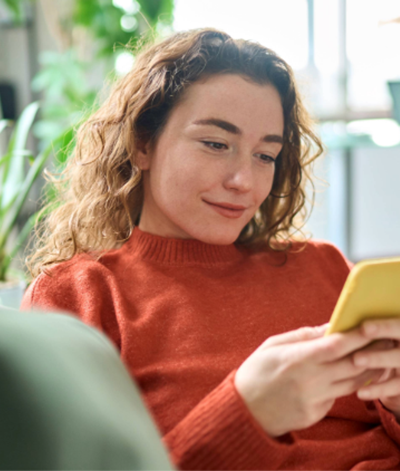 Woman with curly hair wearing an orange sweater, sitting on a sofa, looking at her smartphone in a bright, modern living room.