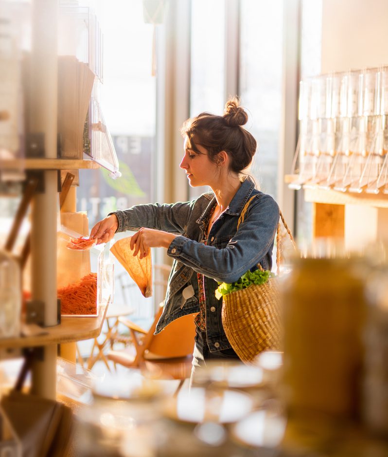 A woman shopping in a bulk food store, holding a paper bag and scooping pasta from a bin, with sunlight streaming through large windows behind her.