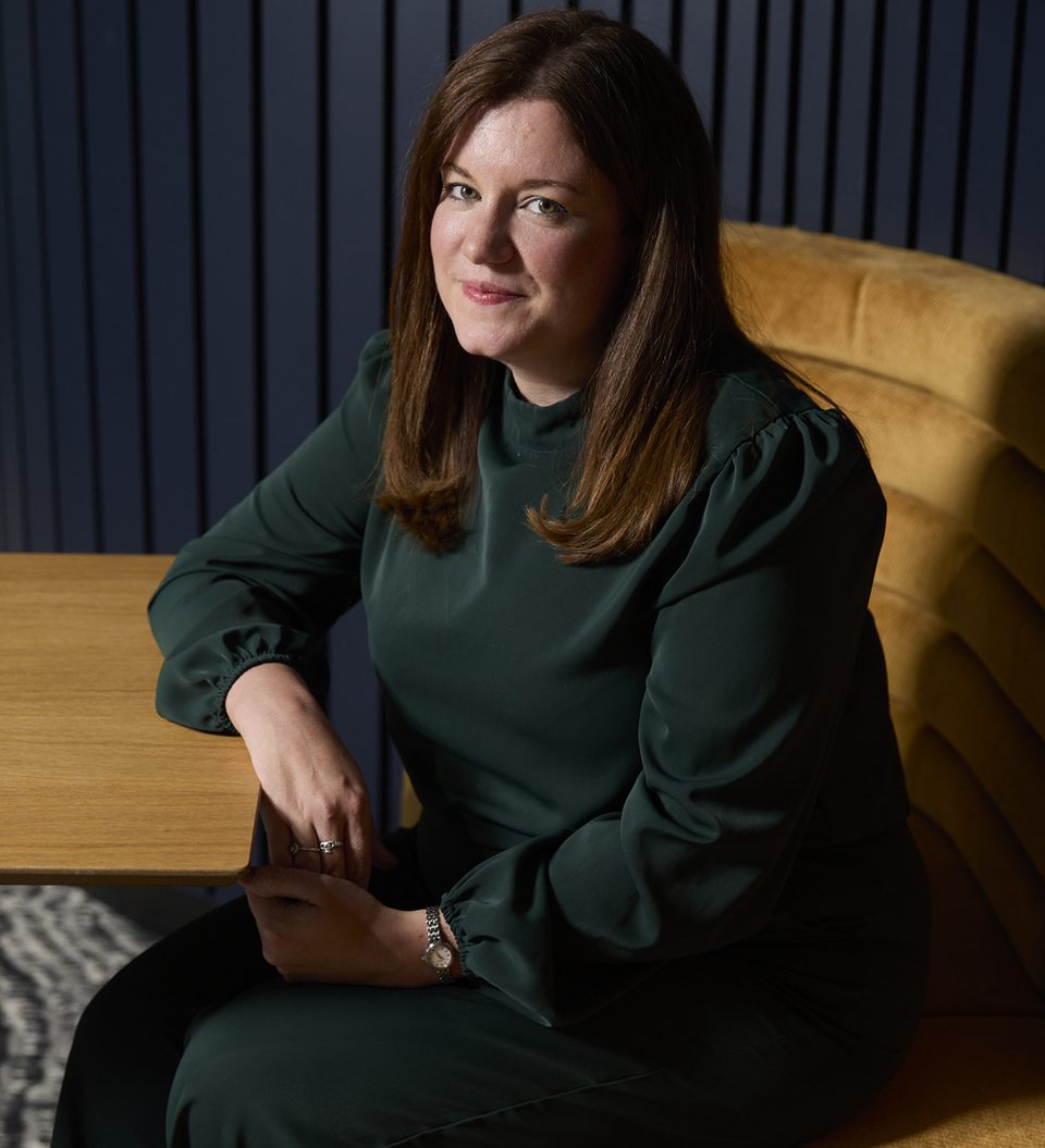 Person with shoulder-length brown hair in a dark green blouse sits at a wooden table beside a mustard-yellow tufted seat, smiling at the camera.