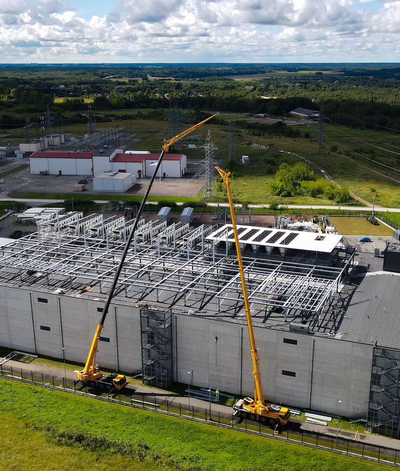 Aerial view of a large industrial complex under construction, with two yellow cranes towering over a steel roof frame, surrounded by fields and power lines.