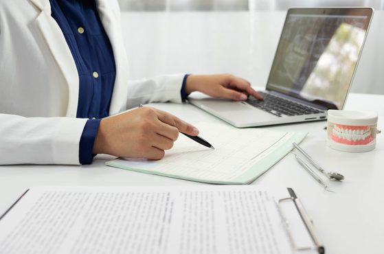 A person in a white coat is at a desk with an open laptop, dental model, and dental tools, pointing at a document with a pen, in a bright clinic setting.
