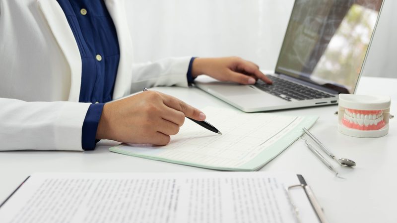 A person in a white coat is at a desk with an open laptop, dental model, and dental tools, pointing at a document with a pen, in a bright clinic setting.