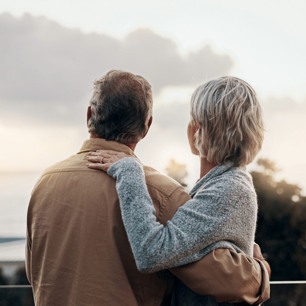An elderly couple stands outdoors, facing a cloudy sky, with the woman resting her hand on the man's shoulder, both looking away from the camera.