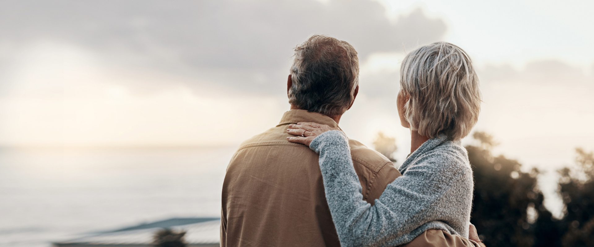 An elderly couple stands outdoors, facing a cloudy sky, with the woman resting her hand on the man's shoulder, both looking away from the camera.