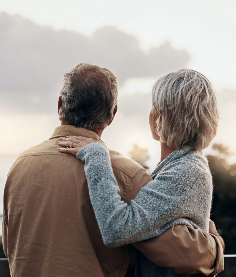 An elderly couple stands outdoors, facing a cloudy sky, with the woman resting her hand on the man's shoulder, both looking away from the camera.