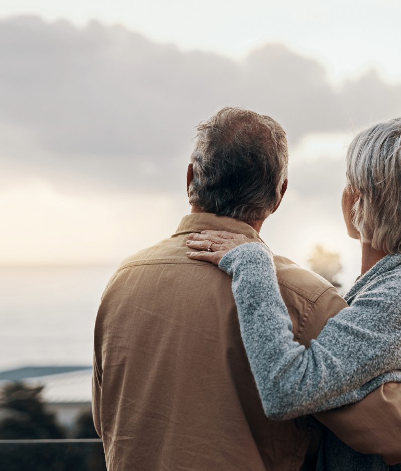 An elderly couple stands outdoors, facing a cloudy sky, with the woman resting her hand on the man's shoulder, both looking away from the camera.