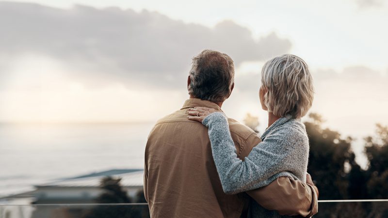An elderly couple stands outdoors, facing a cloudy sky, with the woman resting her hand on the man's shoulder, both looking away from the camera.