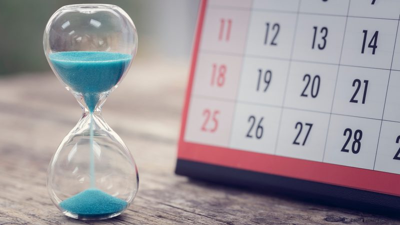 A close-up of a glass timer with blue sand on a wooden surface, with part of a red and white calendar visible in the background.