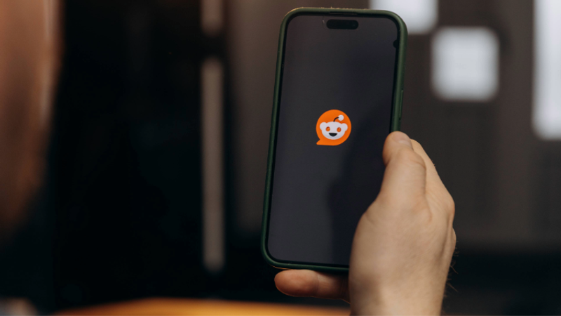 A person holding a phone displaying the Reddit mascot icon on the screen.
