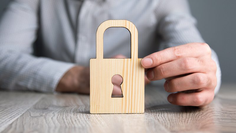 A person in a grey checked shirt holding a wooden padlock-shaped cutout with a keyhole, against a neutral background.