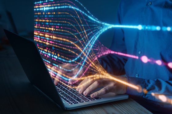 A person using a laptop with colourful digital data streams and waveforms emanating from the screen, representing data transfer or digital communication.