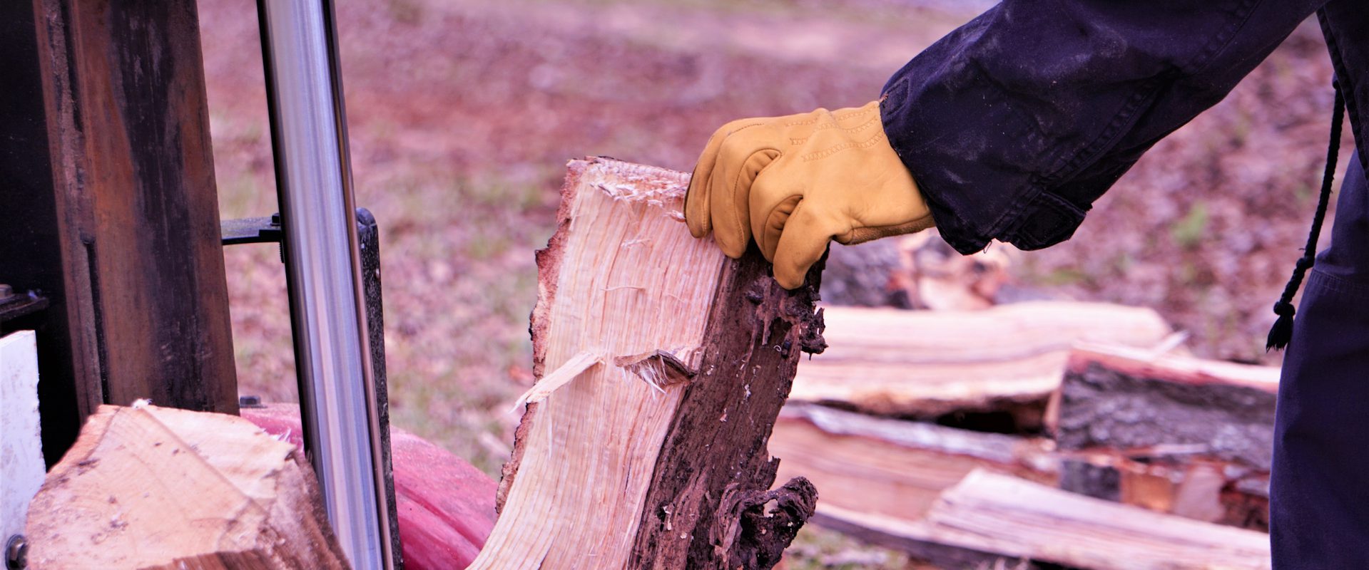 Person splitting firewood with an axe, wearing a black jacket and yellow gloves, outdoors on a ground covered with chopped wood and leaves.