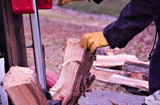 Person splitting firewood with an axe, wearing a black jacket and yellow gloves, outdoors on a ground covered with chopped wood and leaves.
