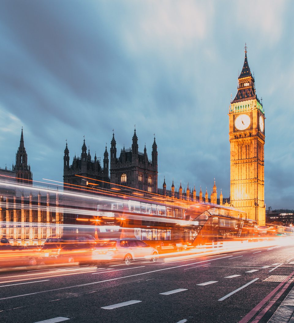 The Palace of Westminster and Big Ben clock tower illuminated at dusk, with light trails from passing vehicles on the busy street in central London.