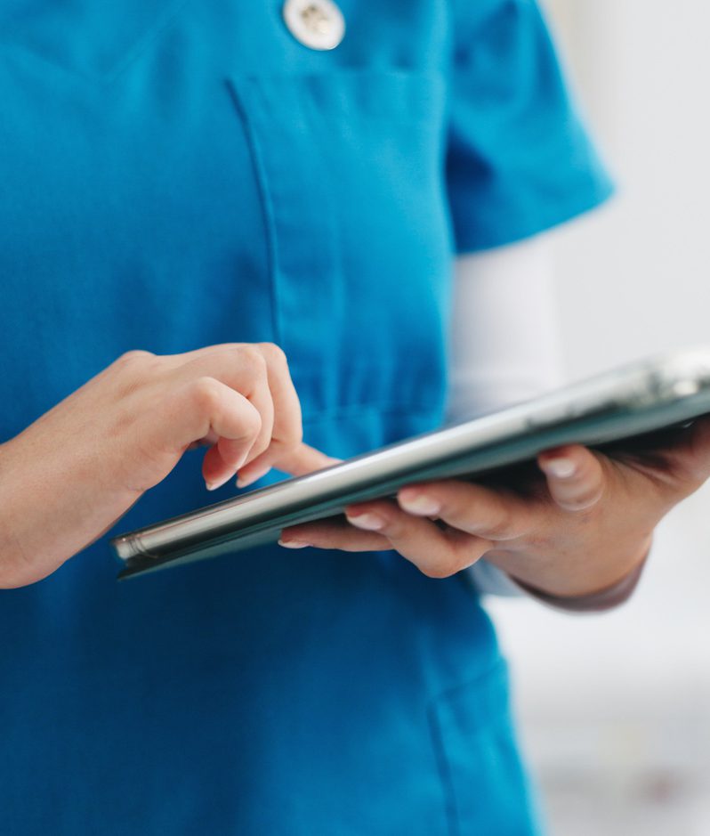 Medical professional in blue scrubs types on a tablet, a stethoscope hanging from the neck; white sleeves show under the top in a bright clinical setting.