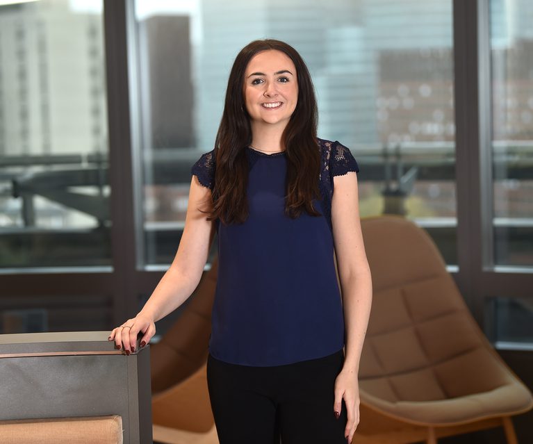 A woman with long brown hair, wearing a navy blue top with lace details on the shoulders, standing in a modern office with large windows and a brown lounge chair.