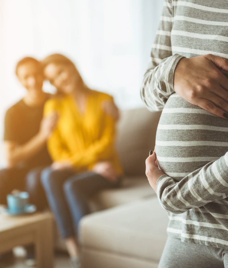 Expectant mother holds stomach with happy couple in background