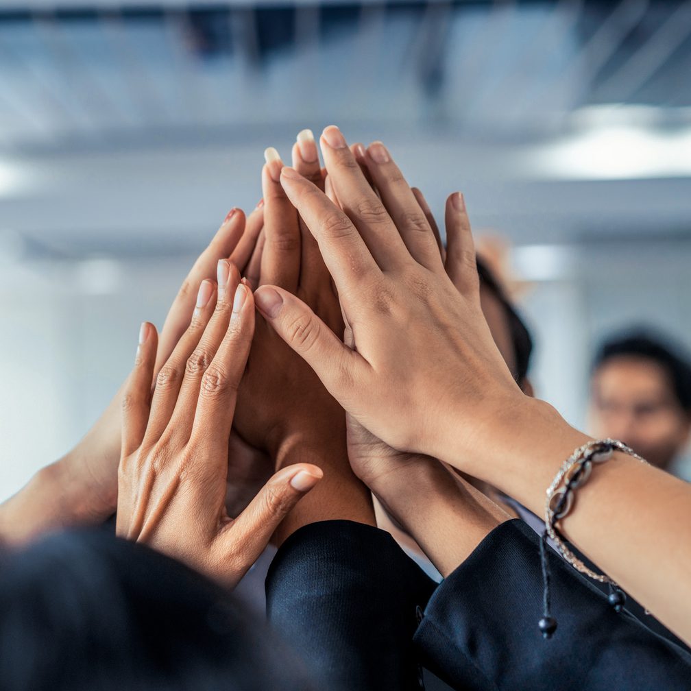 Multiple hands are stacked together in a gesture of teamwork or unity, with blurred individuals in the background in an indoor setting.