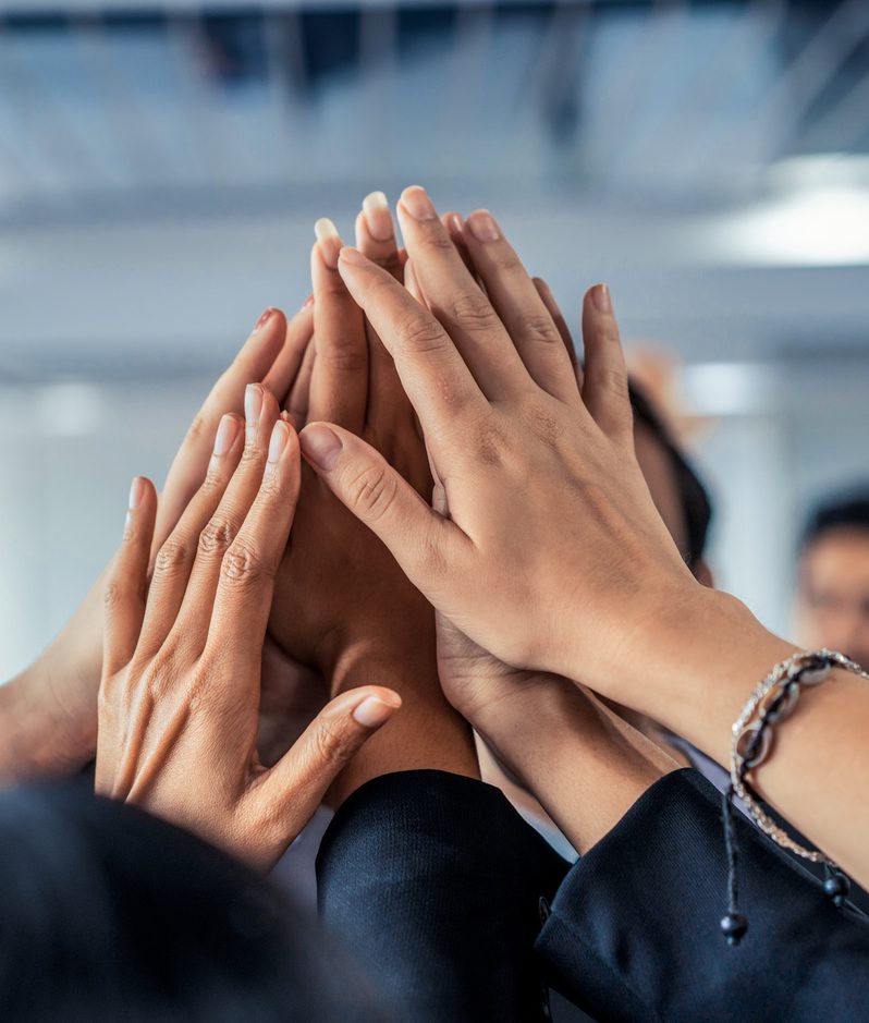 Multiple hands are stacked together in a gesture of teamwork or unity, with blurred individuals in the background in an indoor setting.