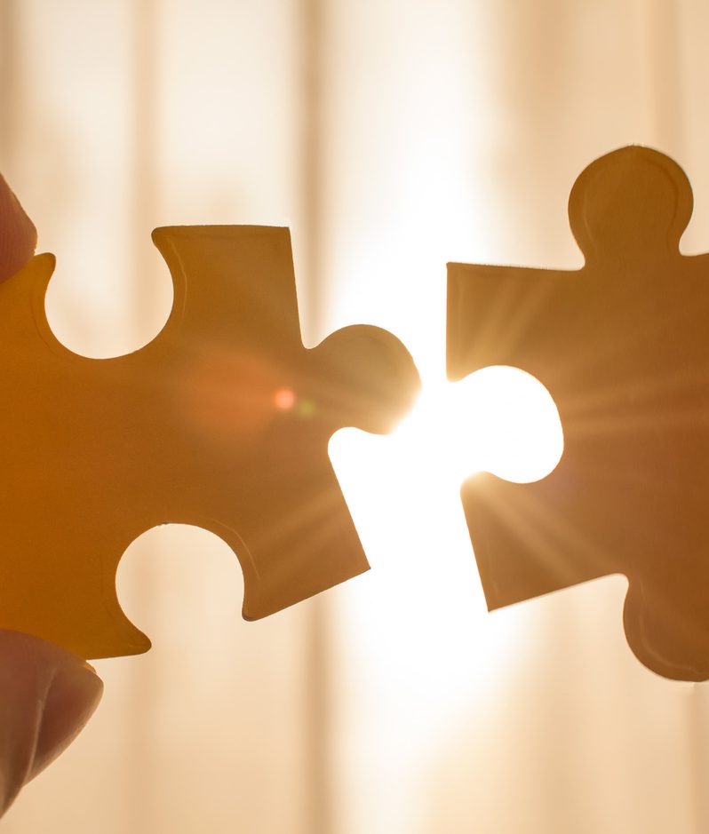 Two hands holding puzzle pieces with the sunlight shining through the gaps between them.