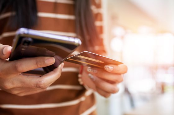 A person holding a smartphone in one hand and a credit card in the other, with sunlight brightening the scene indoors.