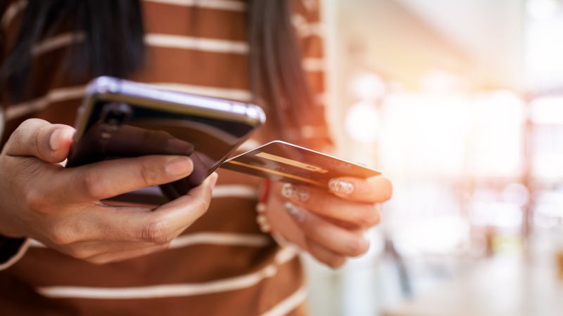 A person holding a smartphone in one hand and a credit card in the other, with sunlight brightening the scene indoors.