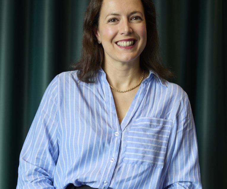 Smiling person with shoulder-length brown hair wearing a light blue striped shirt and a gold chain, seated at a dark table with green curtains in the background.