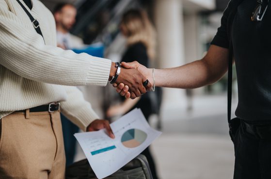 Close-up of two people shaking hands, one holding a printed chart with a pie graph, in a professional setting with other blurred individuals in the background.