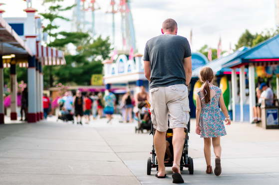 An adult pushes a stroller while a young child walks beside them at a colourful fairground with stalls, rides and other visitors in the background.