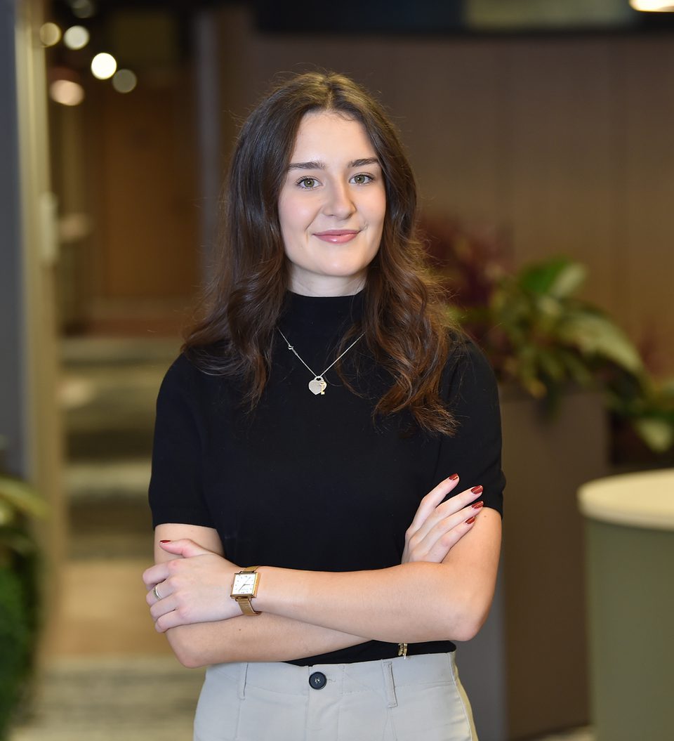 A young woman with long wavy brown hair, wearing a black turtleneck, beige trousers, a necklace with a pendant, and a watch, stands with her arms crossed indoors.