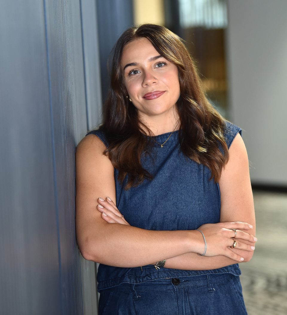 A woman with long brown hair, wearing a denim sleeveless top, stands against a blue wall with arms crossed, smiling softly in an indoor setting.